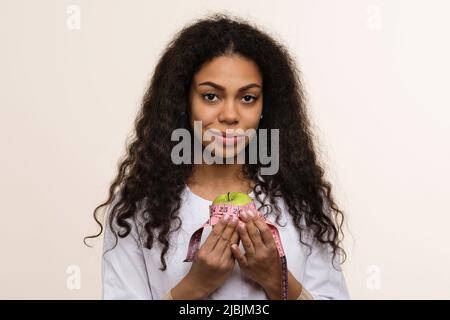 Nahaufnahme Porträt Afrikanisch-amerikanische Ernährungswissenschaftlerin. Freundlich Dunkle Haut in Uniform hält in den Händen Grüner Apfel und Messband auf hellem Backgr Stockfoto