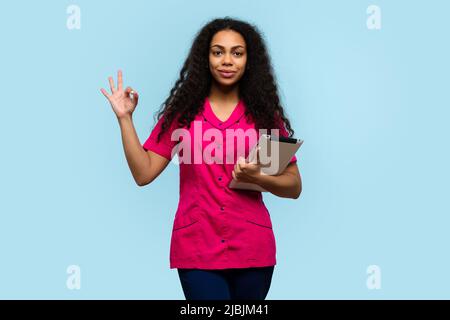 Dunkelhäutige Krankenschwester mit digitalem Tablet. Technologien in der Medizin. Arzt in rosa Uniform zeigt OK-Zeichen auf blauem Hintergrund Stockfoto