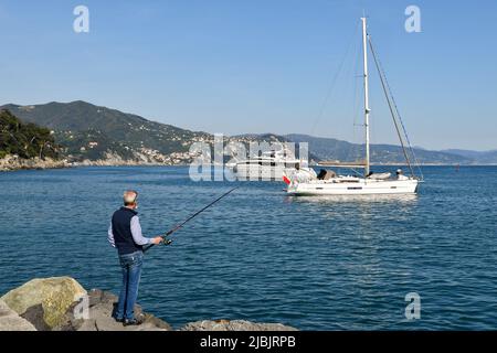 Pensionierte Mann Stange Angeln auf den Felsen des Meeres Dorf mit Luxusbooten vor dem Ufer und der Küste im Hintergrund, Santa Marghertia Ligure Stockfoto