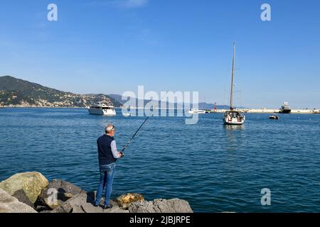 Pensionierte Mann Stange Angeln auf den Felsen des Meeres Dorf mit Luxusbooten vor dem Ufer und der Küste im Hintergrund, Santa Marghertia Ligure Stockfoto