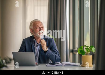 Senior Professional mit einem Notebook, das wegschaut. Ein nachdenklicher Geschäftsmann sitzt am Schreibtisch. Er trägt Formalien in der Unternehmenszentrale. Stockfoto