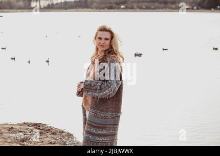 Glückliche, schöne blonde Frau in warmen Kleidern, die allein am Ufer des Flusses an einem kühlen, sonnigen Tag spazieren geht, sich entspannt und genießt, Enten, die im Wasser schwimmen Stockfoto