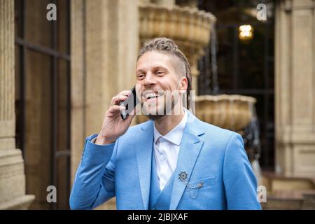 Ein junger Mann mit einer Hipster-Frisur im Business-Anzug spricht fröhlich am Telefon Stockfoto