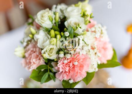 Weicher Brautstrauß aus frischen Blumen und Hochzeit Goldringe auf festlichen Tisch Nahaufnahme, verschwommener Hintergrund. Elegantes Attribut und Tradition Stockfoto