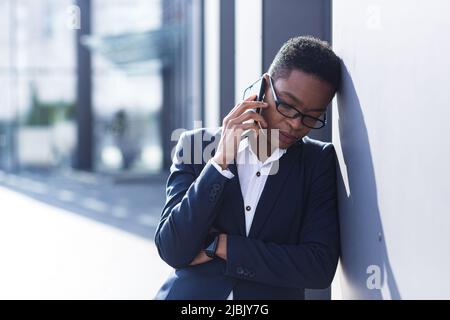 Frau Geschäftsmann traurig beschwert sich am Telefon, berichtet schlechte Nachrichten, afroamerikanische Frau in Verzweiflung in der Nähe des Büros, die Hände hinter seinem Kopf Stockfoto