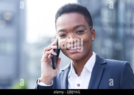 Nahaufnahme eines Porträts einer jungen Geschäftsfrau, einer afroamerikanischen Frau, die am Telefon telefoniert, glücklich und glücklich lächelt, in der Nähe des Büros vor der Tür Stockfoto
