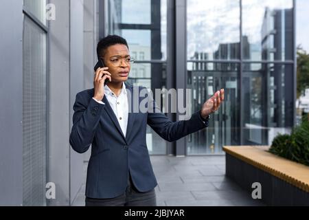 Seriöse und nervöse Geschäftsfrau mit Brille, afroamerikanische Frau mit kurzem Haarschnitt, die am Telefon telefoniert, außerhalb des Büros Stockfoto