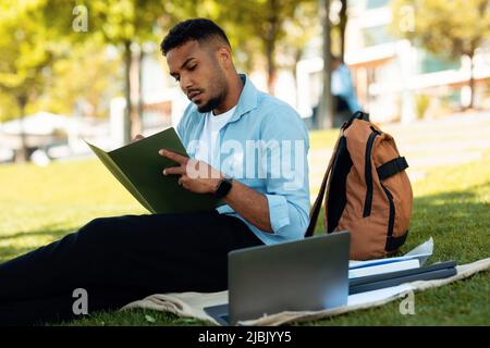 Fokussierter schwarzer männlicher Student, der lernt, Laptop benutzt und Notizen macht, im Park im Freien sitzt. Online-Bildungskonzept Stockfoto