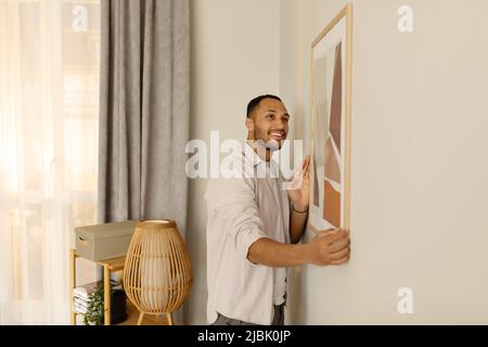 Lächelnder junger schwarzer Mann, der Bilderrahmen setzt und Gemälde an die Wand hängt. Inneneinrichtung und inländische Einrichtung Stockfoto