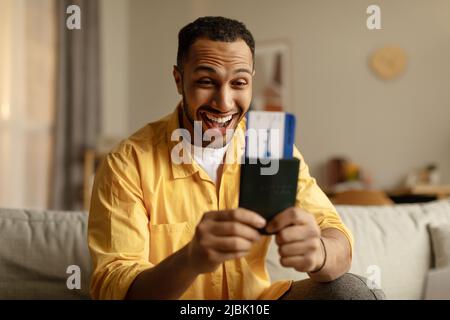 Aufgeregter junger schwarzer Mann mit Pass und Tickets, der sich für den Sommerurlaub vorbereitet und zu Hause auf der Couch sitzt Stockfoto