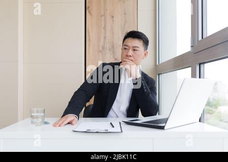 Junger asiatischer Büroangestellter, der bei der Arbeit im Büro an Kopfschmerzen leidet, trinkt Medikamentenpille mit Wasser. Kranker Mann in einem Anzug am Computer in innenräumen wi Stockfoto