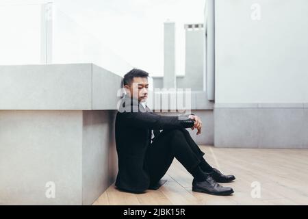 Junger, depressiver Mann, der auf einer Straße im Freien in der Nähe des Bürogebäudes sitzt. Der verärgerte männliche Geschäftsmann verlor seinen Job aufgrund der Finanzia Stockfoto