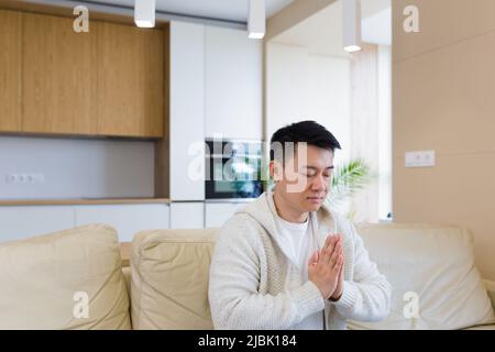 Junger asiatischer Mann, der allein zu Hause betet. Mann faltete Bleistiftarme und meditierte im Apartment-Zimmer. Christliche Religion traditionelle spirituelle für ihre h Stockfoto