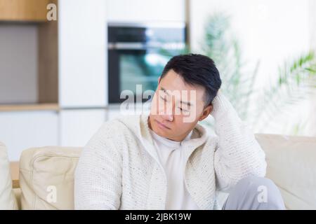 Gelangweilte Freunde junger asiatischer Mann, der allein zu Hause auf dem Sofa sitzt. Vermisst Mann sitzt auf der Couch in der Wohnung. Einsamer Kerl im Wohnzimmer. Ich hatte mir Sorgen gemacht Stockfoto
