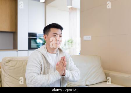 Junger asiatischer Mann, der allein zu Hause betet. Mann faltete Bleistiftarme und meditierte im Apartment-Zimmer. Christliche Religion traditionelle spirituelle für ihre h Stockfoto