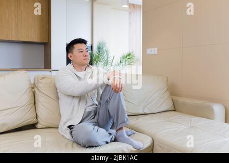 Gelangweilte Freunde junger asiatischer Mann, der allein zu Hause auf dem Sofa sitzt. Vermisst Mann sitzt auf der Couch in der Wohnung. Einsamer Kerl im Wohnzimmer. Ich hatte mir Sorgen gemacht Stockfoto