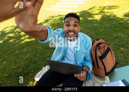 Aufgeregt schwarzen männlichen Studenten geben High Five an Freund, während mit Laptop-Computer, sitzen auf Gras im Park draußen Stockfoto