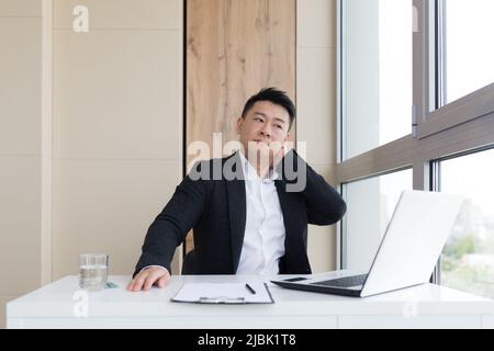 Junger asiatischer Büroangestellter, der bei der Arbeit im Büro an Kopfschmerzen leidet, trinkt Medikamentenpille mit Wasser. Kranker Mann in einem Anzug am Computer in innenräumen wi Stockfoto