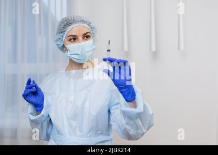 Porträt einer jungen Ärztin Krankenschwester in weißem Mantel in Spaltmaske OP-Kappe und blauen medizinischen Handschuhen halten Spritze, Blick auf die Kamera Stockfoto