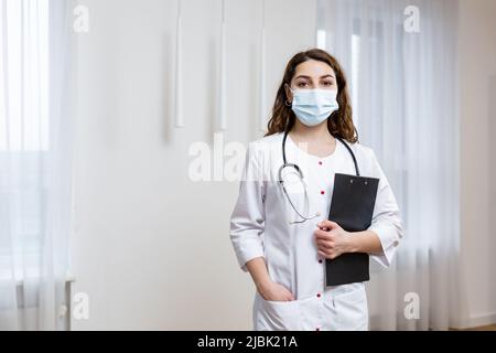 Porträt einer jungen Ärztin in medizinischer Maske im weißen Mantel mit Stethoskop, Ordner mit Dokumenten haltend, Kamera anblickend, Quarantäne Stockfoto