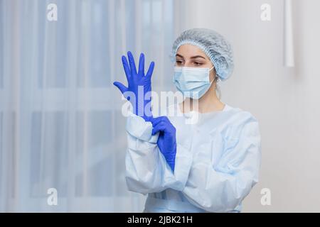Junge Ärztin bereitet sich auf die Operation vor, trägt blaue OP-Handschuhe, in weißem Mantel und Maske, schaut auf die Kamera Stockfoto