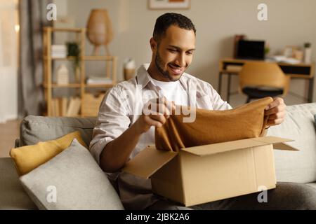 Zufriedener afroamerikanischer männlicher Kunde, der neue Kleidung aus dem Karton herausnimmt, zufrieden mit dem Lieferservice zu Hause Stockfoto