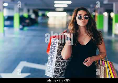 Junge schöne glückliche Frau geht in der Unterführung des Einkaufszentrums zu ihrem Auto mit Einkaufstaschen, Geschenken, Shopaholic Stockfoto