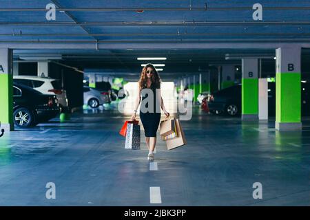 Junge schöne glückliche Frau geht in der Unterführung des Einkaufszentrums zu ihrem Auto mit Einkaufstaschen, Geschenken, Shopaholic Stockfoto