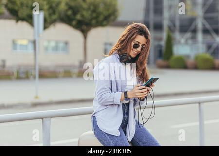 Junge schöne Frau, die mit einem Koffer, einem Telefon und Kopfhörern auf der Straße sitzt, Musik hört, ruft, wartet Stockfoto