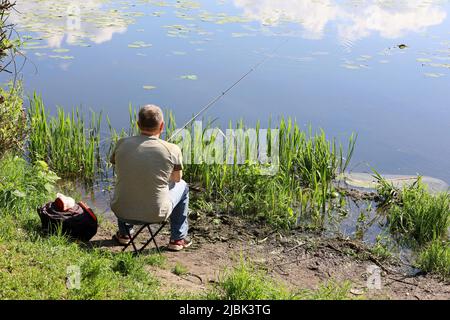Fischer, der mit einer Angelrute am Wasser sitzt, Rückansicht. Mann angeln an der Seenküste im Sommer Stockfoto