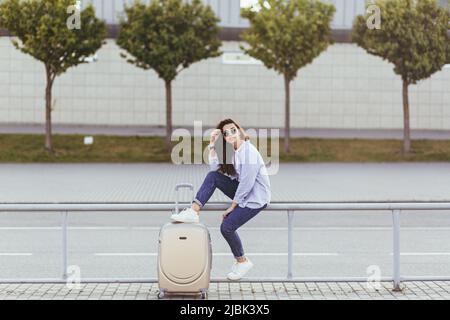 Junge Frau mit einem Koffer auf der Straße in der Nähe des Flughafens, Bahnhof, wartet an der Bushaltestelle, wartet auf ein Taxi Stockfoto