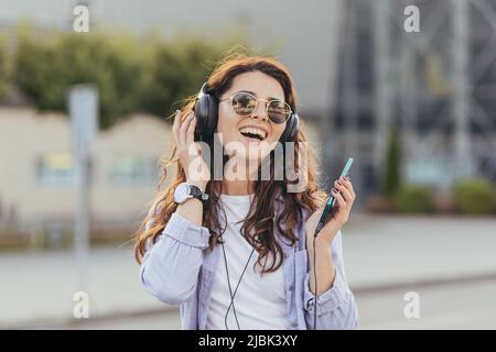 Porträt einer jungen schönen Frau, die fröhlich Musik vom Telefon in Kopfhörern hört, auf der Straße sitzend, in der Nähe des Flughafens, des Bahnhofs, Stockfoto