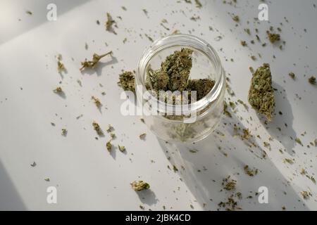 Unkrautladen. Glas mit getrockneten Cannabisknospen auf weißem Hintergrund mit zerdrücktem Marihuana. Stockfoto