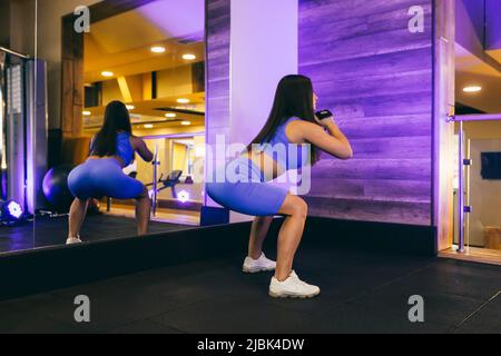 Junge schöne Sportfrau im Fitnessstudio macht Übungen, Kniebeugen, vor dem Spiegel, in Sportuniform Stockfoto