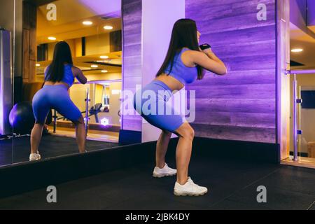 Junge schöne Sportfrau im Fitnessstudio macht Übungen, Kniebeugen, vor dem Spiegel, in Sportuniform Stockfoto