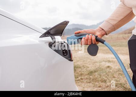 Männliche Hand, die ein Ladegerät in die Steckdose für Elektroautos steckt, im Hintergrund sichtbare Bergnatur Stockfoto