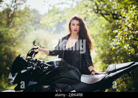 Porträt einer schönen jungen Frau auf einem Motorrad in schwarzer Lederkleidung auf der Natur, Straße, Wald, bei sonnigem Wetter, Nahaufnahme, Betrachten Sie die Stockfoto