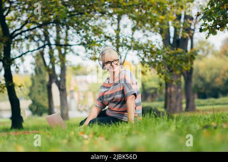 Schöne blonde Frau, Senior, sitzt auf dem Gras im Park in Kopfhörern, nutzt einen Laptop, arbeitet, studiert, hört Musik, Vorträge auf dem Video pl Stockfoto