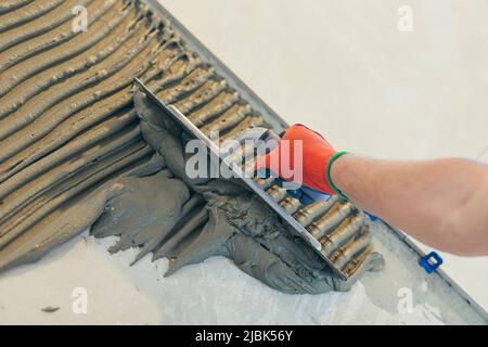 Nahaufnahme der Hände eines Bauherrn mit einer gekerbten Kelle, setzen eine Fliese Klebstoff-Mischung, führt die Installation von Keramikfliesen Stockfoto