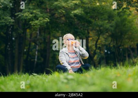 Ältere schöne Frau sitzt im Park auf dem Gras und trinkt frischen Saft, Wasser nach dem Sport, Yoga Stockfoto