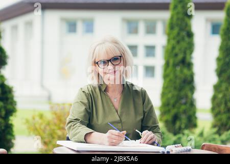Ältere schöne blonde Frau sitzt an einem Tisch auf der Straße, malt ein Bild, lächelnd, glücklich Stockfoto
