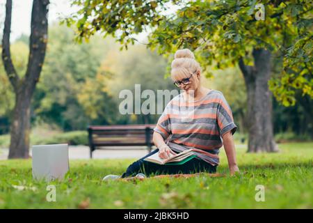 Ältere schöne blonde Frau, die im Park auf dem Gras sitzt, liest ein Buch, studiert, lächelt, ruht Stockfoto