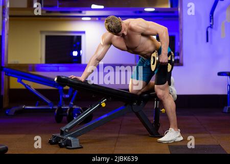 Junger, hübscher, athletischer Mann, der Übungen im Fitnessstudio mit Hanteln macht, in vollem Wachstum, mit Blick auf die Kamera Stockfoto