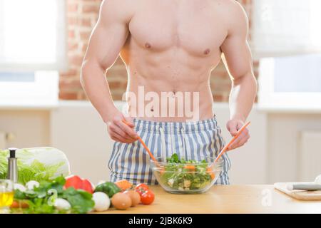 Porträt eines jungen, hübschen, athletischen Magns, der in der Küche zu Hause einen Salat mit frischem Gemüse zubereitet und die Kamera anschaut, Smiley Stockfoto