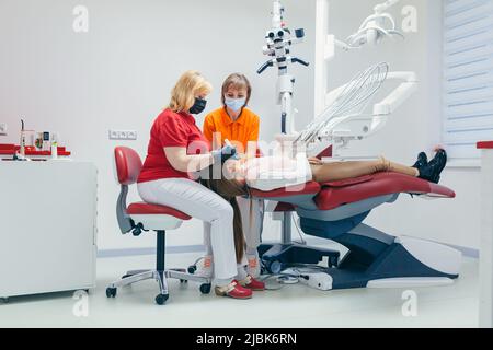 Junge schöne Patientin in der Zahnarztpraxis behandelt Zähne, liegt auf einem Zahnstuhl, Arzt und Assistent behandelt die Zähne des Patienten in Masken, glo Stockfoto