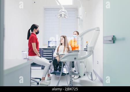 Junge schöne Patientin bei einem Zahnarzttermin, sitzt auf einem Zahnarztstuhl und spricht mit einem Arzt und einer Assistentin, bereitet sich auf die Behandlung vor Stockfoto