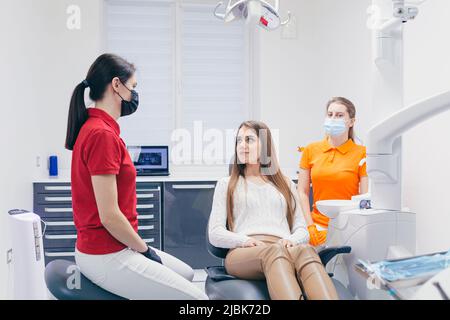 Junge schöne Patientin bei einem Zahnarzttermin, sitzt auf einem Zahnarztstuhl und spricht mit einem Arzt und einer Assistentin, bereitet sich auf die Behandlung vor Stockfoto