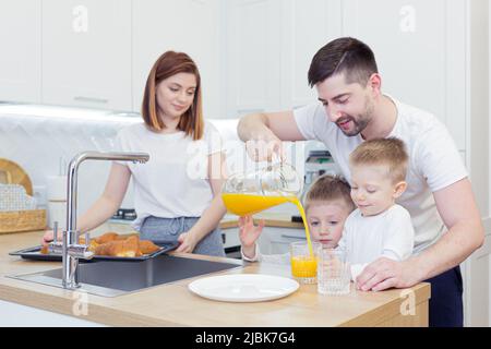 Familienfrühstück, Mama, Papa, zwei Kinder, Jungen, die von Mama gebackene Croissants aßen, Kaffee und frischen Saft tranken, lächelten und sich zu Hause in der Kitch umarmten Stockfoto
