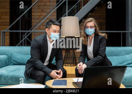Zwei Mitarbeiter eines internationalen Unternehmens diskutieren einen Bericht über die geleistete Arbeit, ein asiatischer Mann und eine Frau, die medizinische Schutzmasken tragen, sitzen im Stockfoto
