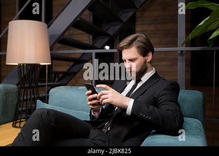Ein männlicher Geschäftsmann sitzt in einem stilvollen Restaurant, entspannt in einem Geschäftsanzug und nutzt das Telefon, um zu kommunizieren Stockfoto